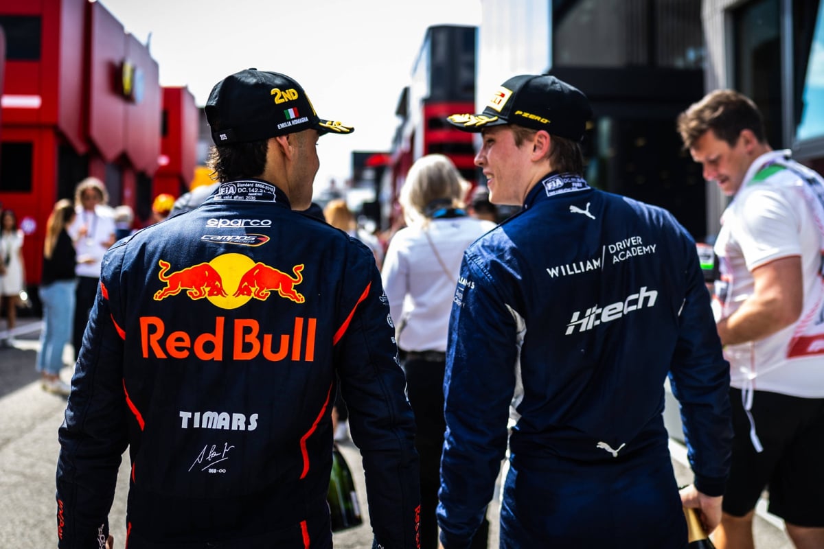 Lindblad and Browning facing each other with their backs to the camera in the paddock wearing Red Bull and Williams Academy F2 race suits