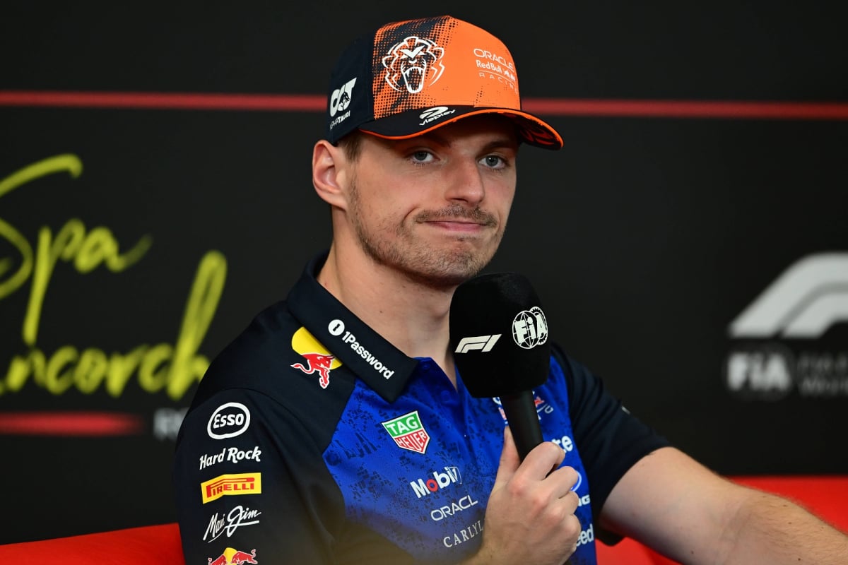 Verstappen in blue Red Bull tee and orange cap holding FIA and F1 mic looking smug