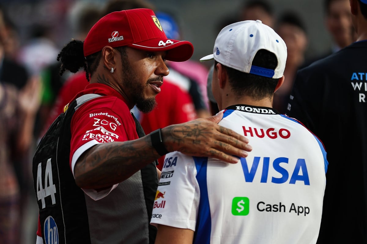 Hamilton in Ferrari kit with his arm around Hadjar, who faces away from the camera in Racing Bulls teamwear in the F1 paddock