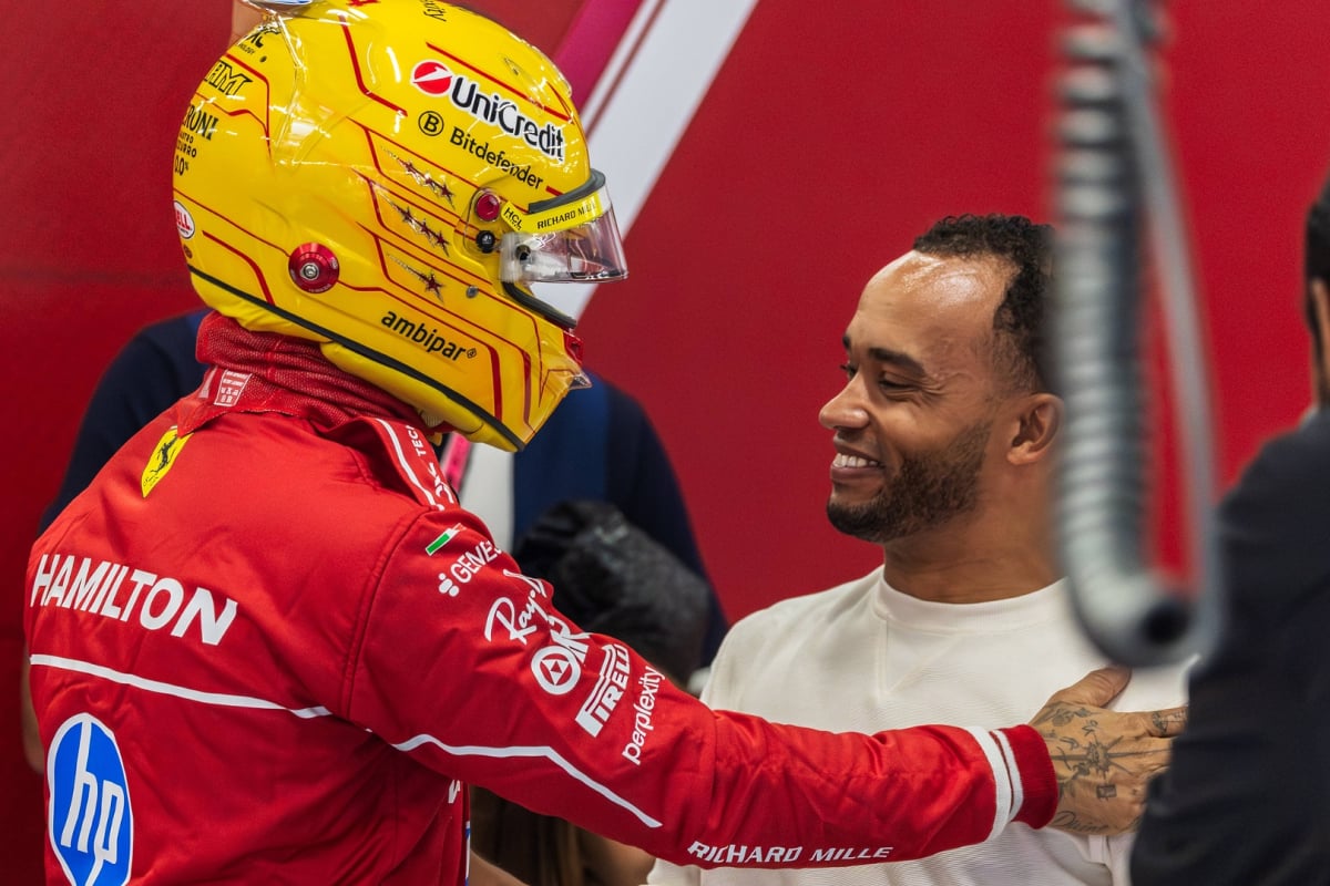 Lewis Hamilton in yellow helmet and red Ferrari care suit with a hand on the shoulder of brother Nicolas who is smiling in Ferrari garage