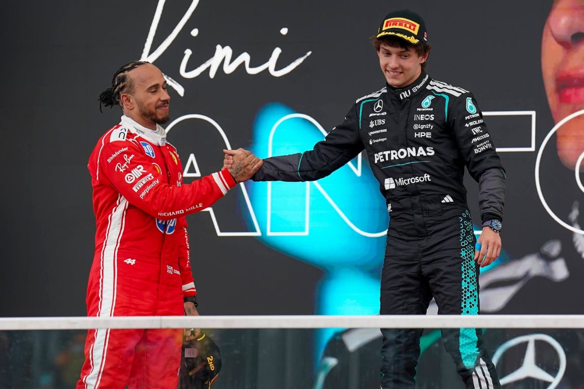 Hamilton on podium below shaking hand of Antonelli who is on top step, both in Ferrari and Mercedes race suits respectively 