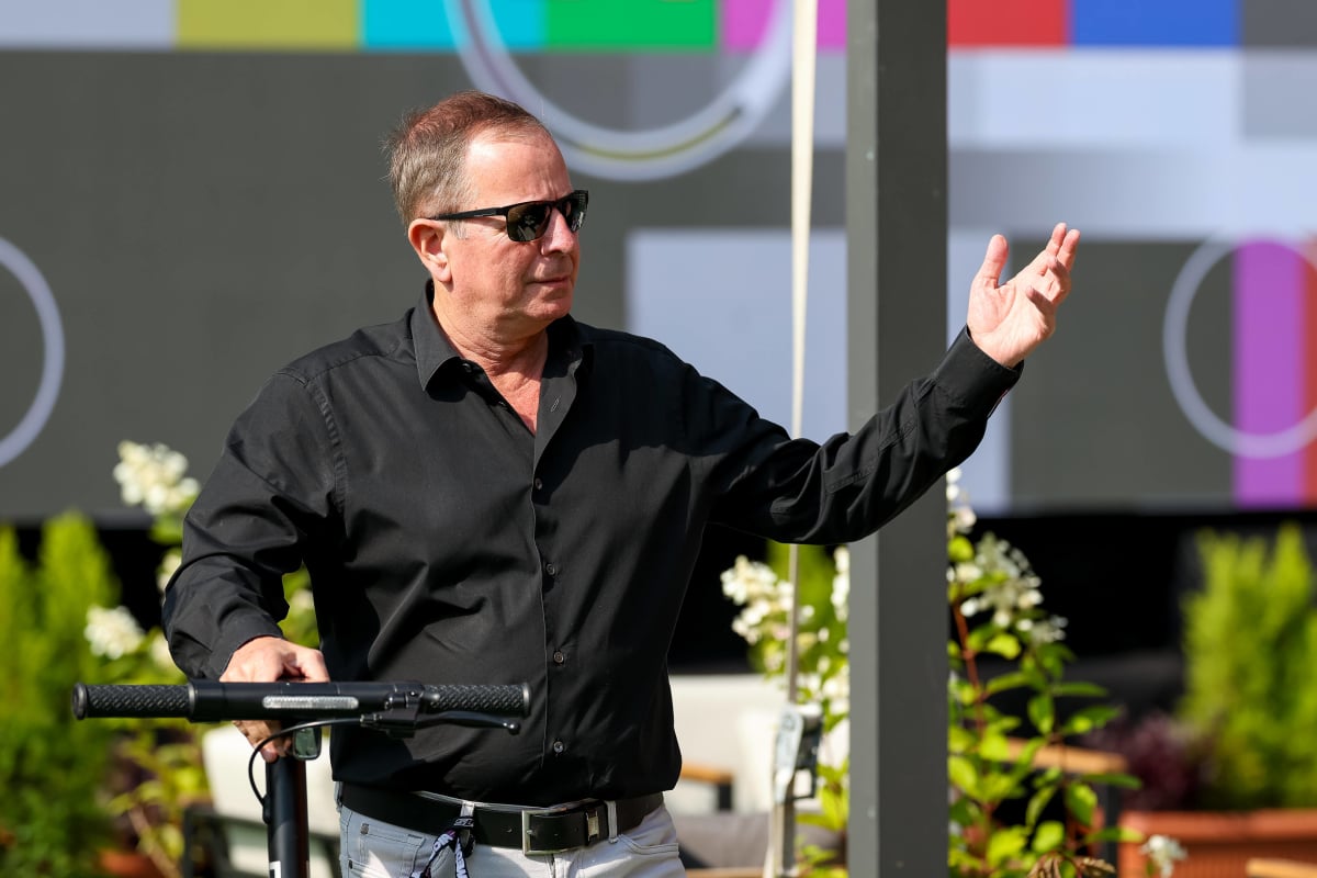 Martin Brundle wheels a bike and gesticulates with his hand looking into the distance, somewhat annoyed