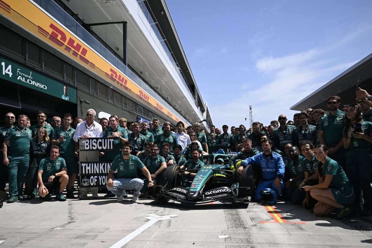 Team picture of Aston Martin F1 team posing around Alonso's car in front of his garage with 'Biscuit thinking of you' sign
