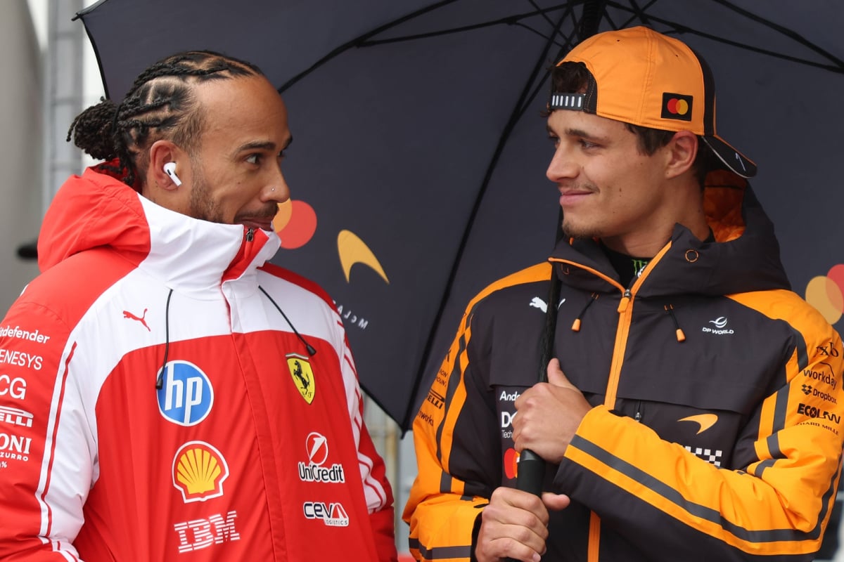Hamilton looking cheekily at Norris who is smiling, stood under the same umbrella in Ferrari and McLaren kit respectively