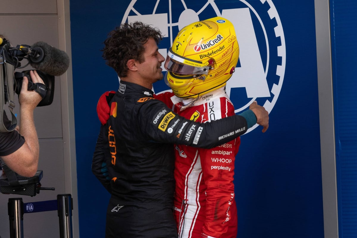 Norris and Hamilton hugging after Barcelona GP in race suits with blue wall in background with white FIA logo on it