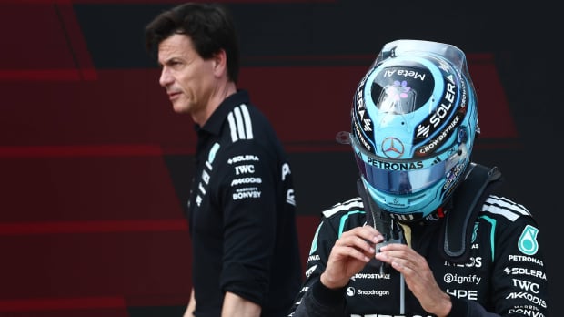 Russell in turquoise F1 helmet with Wolff walking past in the background looking serious — Foto: © IMAGO