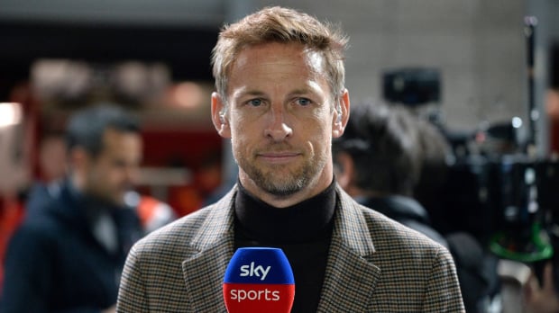 Button in high-neck dark top and beige blazer in the F1 paddock holding a Sky Sports microphone