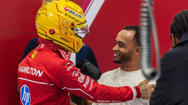 Lewis Hamilton in yellow helmet and red Ferrari care suit with a hand on the shoulder of brother Nicolas who is smiling in Ferrari garage — Photo: © IMAGO