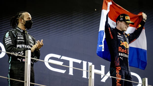 Hamilton on the second step of the Abu Dhabi 2021 podium with a face mask on and Verstappen flying the Dutch flag, smiling on the top step