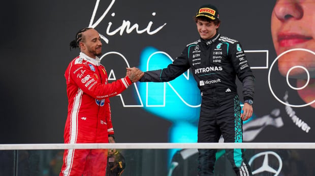 Hamilton on podium below shaking hand of Antonelli who is on top step, both in Ferrari and Mercedes race suits respectively — Foto: © IMAGO