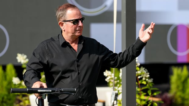 Martin Brundle wheels a bike and gesticulates with his hand looking into the distance, somewhat annoyed
