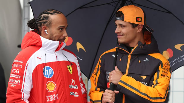Hamilton looking cheekily at Norris who is smiling, stood under the same umbrella in Ferrari and McLaren kit respectively — Photo: © IMAGO