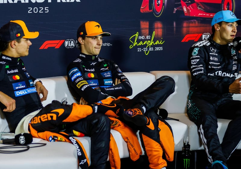Norris and Piastri looking annoyed at Russell who is holding the F1 mic on the press conference sofa