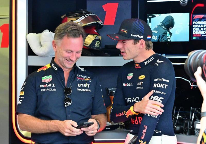 Christian Horner laughs while looking at his phone, while Max Verstappen smiles alongside him