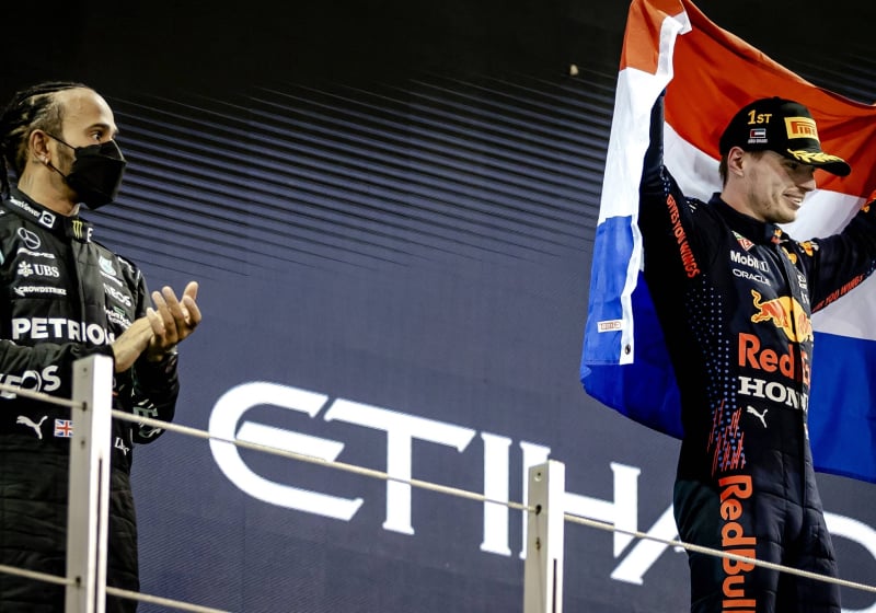 Hamilton on the second step of the Abu Dhabi 2021 podium with a face mask on and Verstappen flying the Dutch flag, smiling on the top step