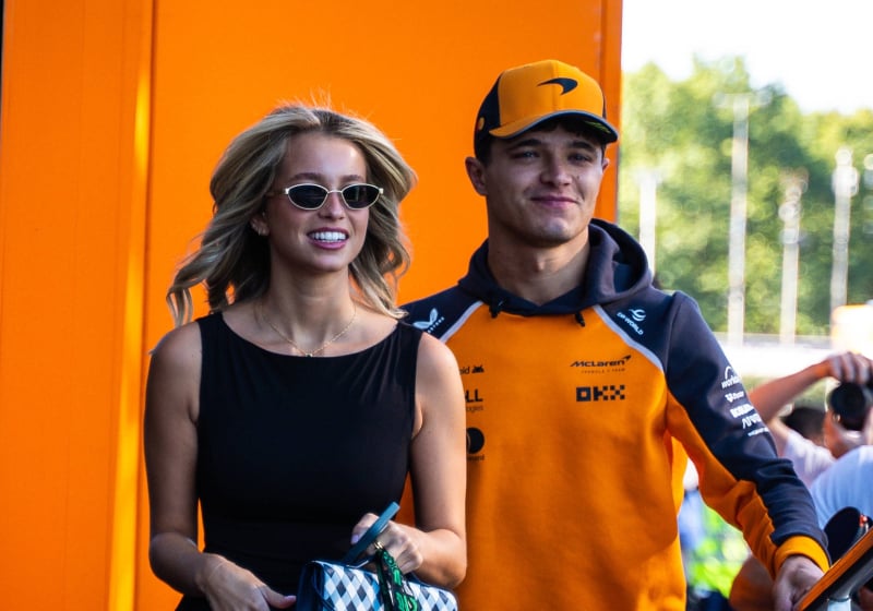 Norris and his girlfriend smiling as they walk into the McLaren motorhome in an F1 paddock